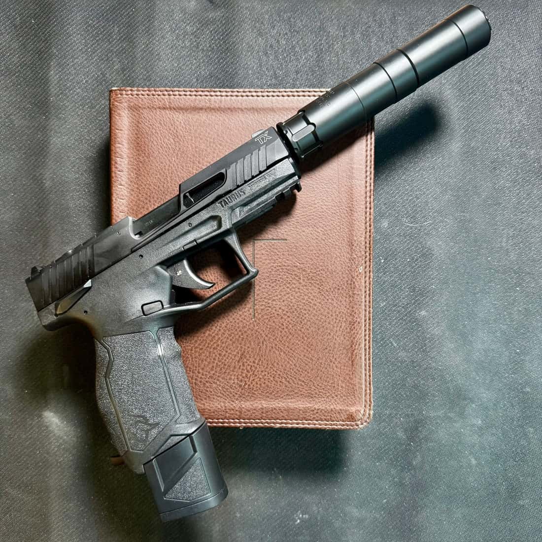 A Rugged Oculus22 suppressor threaded onto a black Taurus TX22 TORO pistol, resting at an angle across a brown leather notebook on grey fabric.