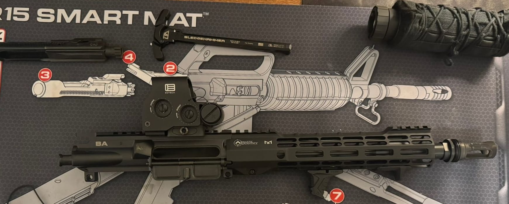 The 10.5 upper broken down on a cleaning mat: bolt carrier group removed and laid alongside, the Breek Arms Sledgehammer charging handle beside it, the ASR flash hider on the muzzle, and the suppressor wearing its cover at the top of the frame.