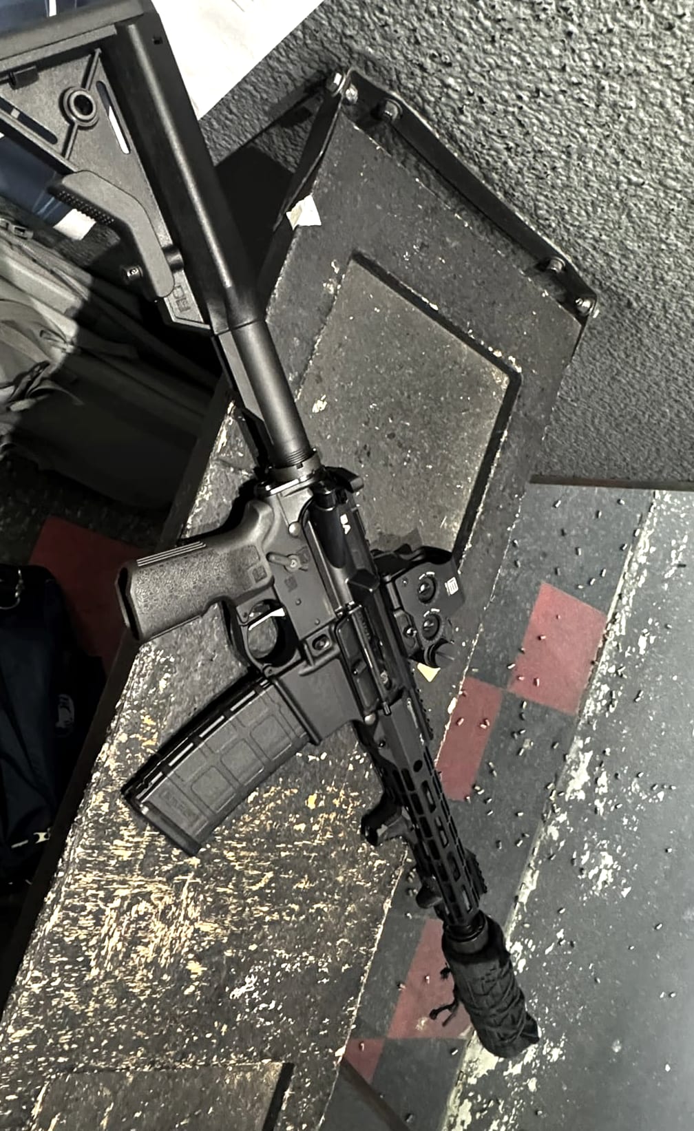 The 10.5 carbine resting on a bench at an indoor range with the Corset cover fitted over the suppressor, spent brass scattered on the floor of the bay.