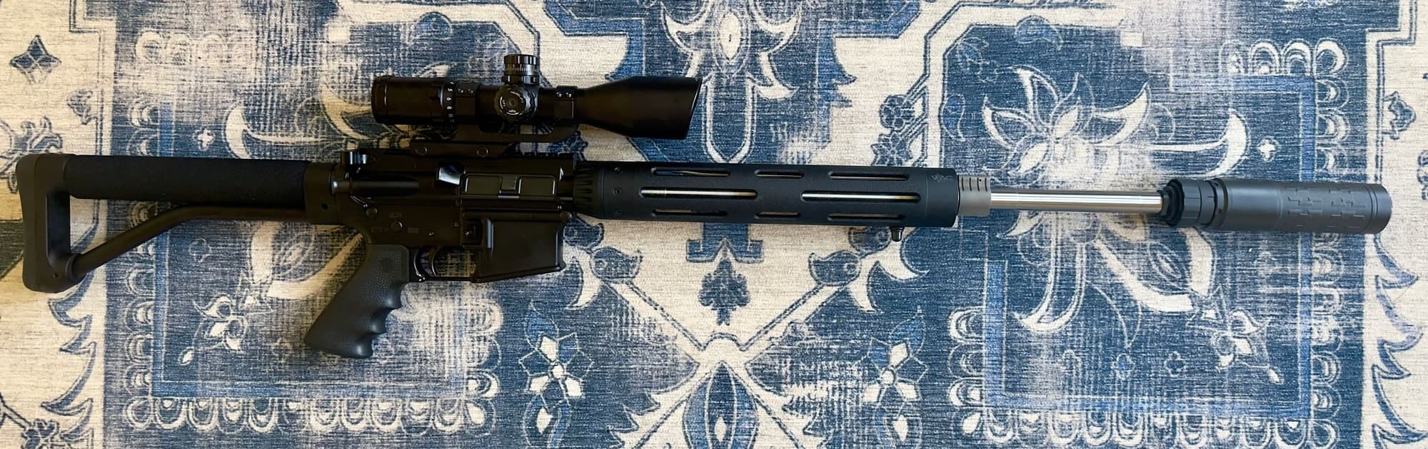 The 20-inch precision build laid out on a patterned rug, showing the full rifle from fixed stock to the Velos suppressor on the muzzle, with a scope on top.