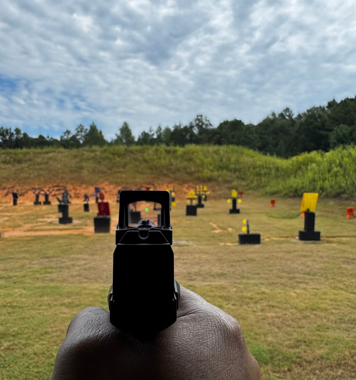 A shooter's-eye view over a pistol fitted with the Vortex Defender-CCW at an outdoor range, the green dot visible in the window, with black and yellow steel targets arranged across the grass in front of a dirt berm.